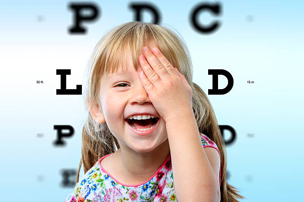 Close up face portrait of happy girl having fun at vision test.Conceptual image with girl closing one eye with hand and block letter eye chart in background.